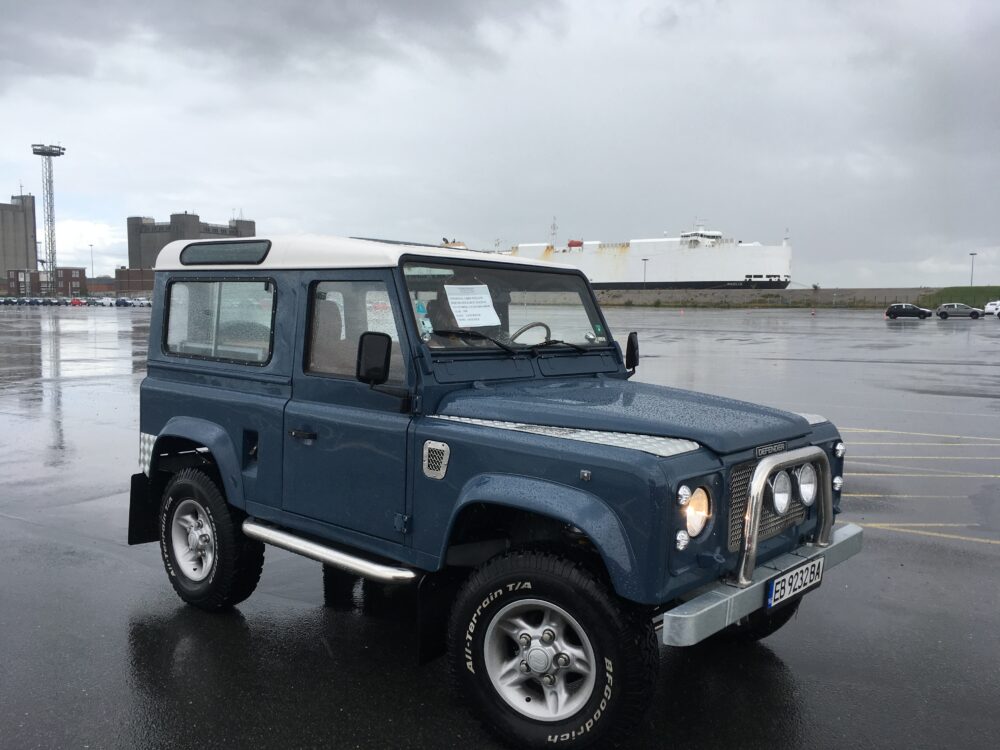 A restored Arles Blue Land Rover Defender 90 waiting at the port, with the ferry behind it, ready to ship