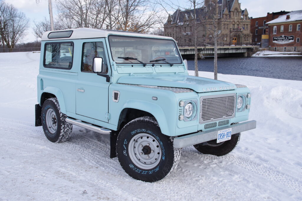 Front three-quarter view of Blue Crush, a matte Sonic Blue Defender 90 with silver Wolf wheels, by a snowy waterfront