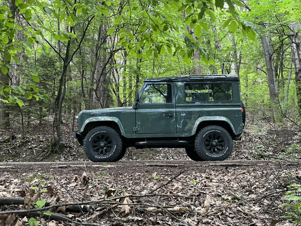 Side profile of The Evergreen, a Keswick Green Defender 90 with a Santorini Black roof, among the trees