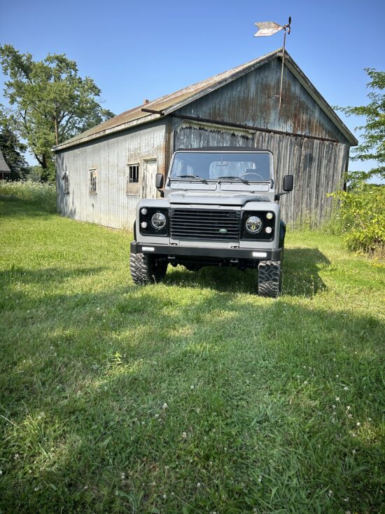 Head-on view of The Nazaré, a Dolphin Grey Land Rover Defender 90 soft top, in front of an old wooden barn